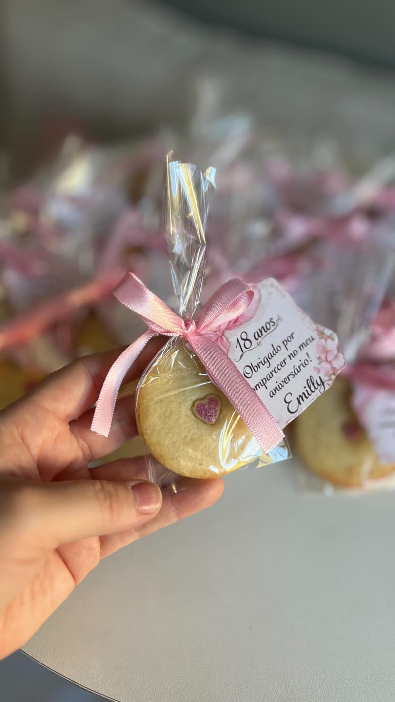 Biscoito decorado embalado com laço rosa e tag impressa com recado de agradecimento, segurado na mão