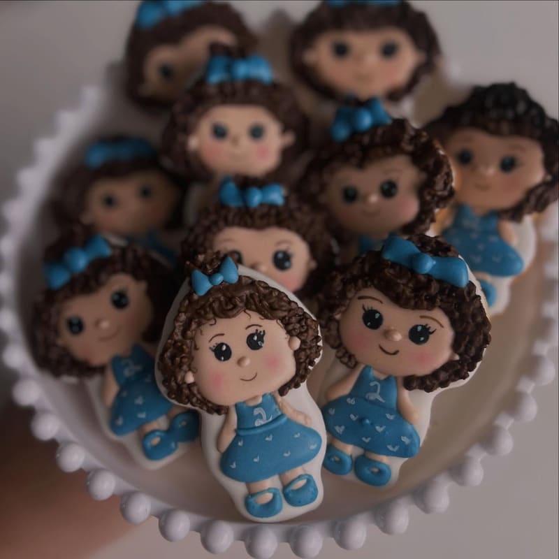 Vários biscoitos decorados em formato de boneca, com laço azul no cabelo, sobre um prato branco